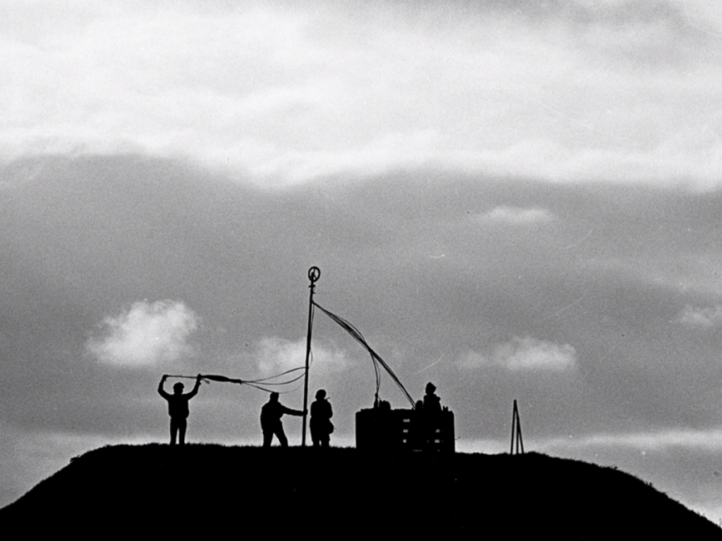 Photo of a group of women dancing around a maypole topped with the peace symbol, on top of a mound, silhouetted against a cloudy sky.