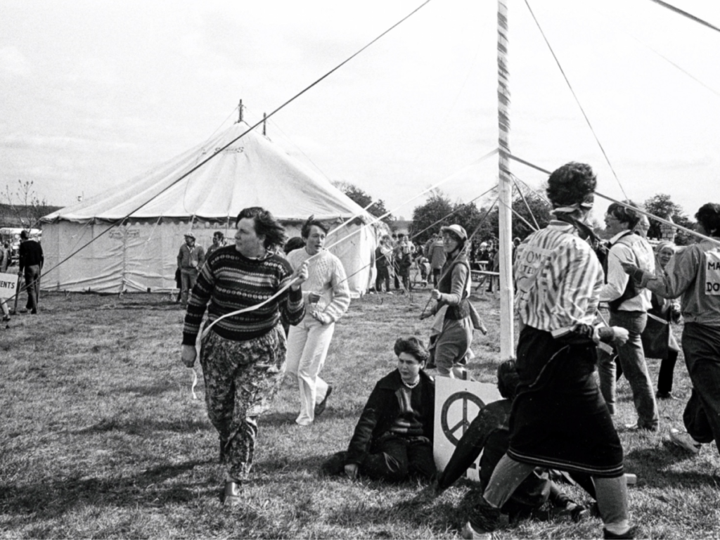 A group of women dancing around a maypole, with a sign with the peace symbol on the ground propped against it. In the background are tents and other activists holding signs.